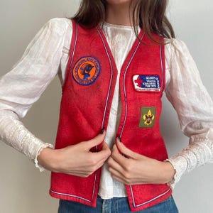 May include: A vintage red vest with white trim and multiple patches. Patches include text that reads "INTERNATIONAL HOUSE OF FRANCE" and "SCOUT-O YOUTH EXPLORES THE 70'S". The vest is open and worn over a white blouse.