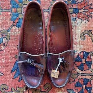 May include: A pair of burgundy leather loafers with tassel details. The shoes have a classic design with a rounded toe and a decorative fringe. The soles are a darker shade of brown. The shoes are displayed on a patterned rug.