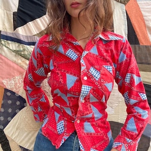 May include: A red button-down shirt with a patchwork design featuring blue and white gingham and triangle patterns. The long-sleeved shirt has a collared neckline and is made of a textured fabric. The shirt is paired with blue jeans.