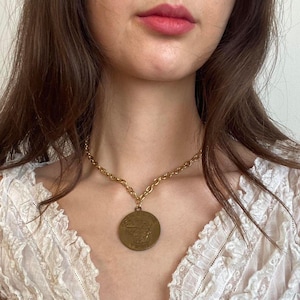May include: A gold-toned necklace with a large, circular pendant. The pendant features a detailed relief design. The chain is a classic link style. The necklace is worn against a white lace top.
