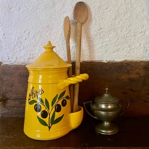 May include: A yellow ceramic olive jar with a lid, featuring a painted olive branch design and the word "olives". Two wooden spoons stand in the jar's side holder, and a small silver container sits nearby. The items are on a wooden surface.