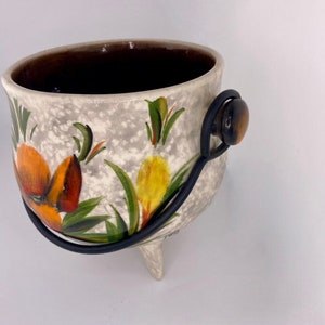 May include: A white ceramic planter with a floral design in shades of orange, yellow, and green. The planter has a black metal handle and three legs.