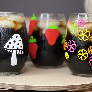 May include: Three clear glass tumblers with black liquid and ice. Each tumbler has a different design: one with white mushrooms, one with red strawberries, and one with colorful flowers.