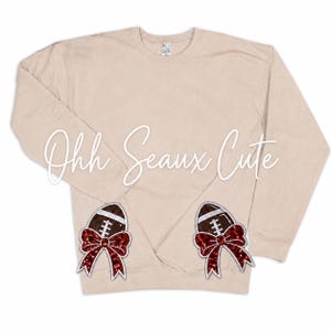 May include: A beige long-sleeve sweatshirt featuring two brown football patches at the bottom, each accented with a large, glittery red bow. The text "Ohh Seaux Cute" is written in white cursive script across the center of the garment.