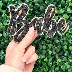May include: A sequined patch in the shape of the word "Babe". The letters are outlined in a cream-colored material and filled with black sequins and a leopard print pattern. The patch is held against a backdrop of green foliage.