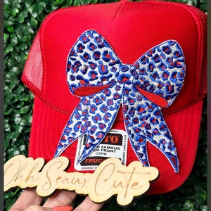 May include: A red trucker hat with a large bow featuring a blue and white leopard print design. The bow is embellished with small white beads. The hat has a mesh back and a curved brim. A wooden sign that says "Ohh Seaux Cute" is in front of the hat.