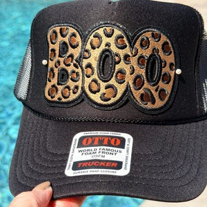 May include: A black trucker hat featuring the word BOO in large, leopard-print letters with black embroidered outlines. The hat has a foam front, a mesh back, and a white sticker on the brim with the text OTTO WORLD FAMOUS FOAM FRONT OSFM TRUCKER.