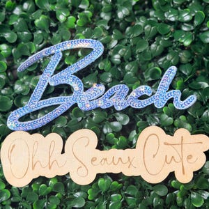 May include: Two decorative signs. The word "Beach" is spelled out in blue sequins. Below it, the phrase "Ohh Seaux Cute" is laser cut from wood. The background is a dense arrangement of green leaves.