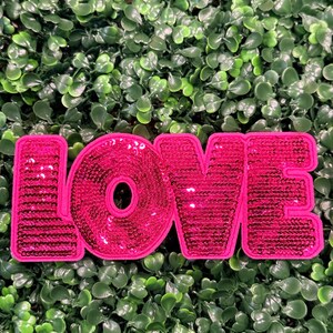 May include: A pink sequined patch spelling out the word "LOVE" against a backdrop of green foliage. The letters are outlined in a darker pink and filled with shiny sequins, creating a textured and eye-catching design.