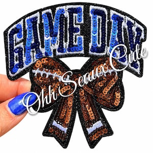May include: A decorative patch featuring the text GAME DAY in blue sequins with white embroidery outlines on a black glitter background, positioned above a brown sequined bow with white embroidered accents and a black glitter border.