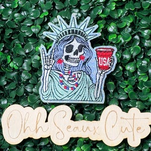 May include: Embroidered patch featuring a skeletal Statue of Liberty holding a coffee cup with "USA" on it. The patch is light blue, red, and white, with a green background. A wooden sign below reads "Ohh Seaux Cute."