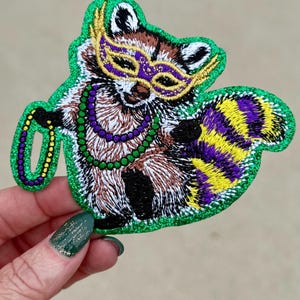 May include: Embroidered patch featuring a raccoon wearing a gold and purple mask and Mardi Gras beads. The raccoon has a striped purple and yellow tail. The patch is outlined in green glitter.