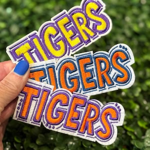 May include: Three embroidered patches with the word "TIGERS" in yellow, orange, and purple. The patches have a white border and are held against a green leafy background. The patches are likely for apparel or accessories.