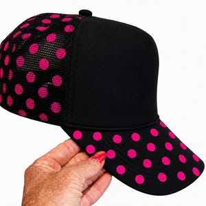 May include: A black baseball cap featuring a solid black front panel and a mesh back panel. The mesh back and the curved brim are decorated with a vibrant hot pink polka dot pattern. The cap has a structured crown and a button on the top center.