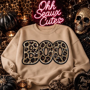 May include: A beige crewneck sweatshirt featuring the word BOO in large, block-style letters. Each letter is filled with a gold and black leopard print pattern and outlined in a thick black border. The fabric has a smooth, solid finish.