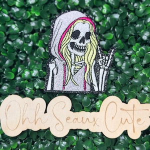 May include: An embroidered patch featuring a skeleton with long blonde hair wearing a grey hoodie with pink trim, making a rock and roll hand gesture. Below the patch is a light-colored wooden cutout with the text Ohh Seaux Cute in cursive.