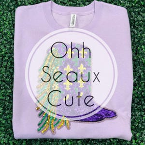 May include: A light purple sweatshirt with a circular graphic. The graphic features a sequined boot design with green, gold, and purple accents, and the words "Ohh Seaux Cute". The sweatshirt is made of a soft material.