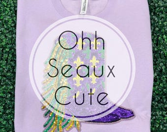 May include: A light purple sweatshirt with a circular graphic. The graphic features a sequined boot design with green, gold, and purple accents, and the words "Ohh Seaux Cute". The sweatshirt is made of a soft material.