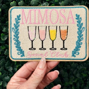 May include: A rectangular patch with the word "MIMOSA" in pink script above four champagne glasses filled with different colored liquids. The text "Social Club" is below the glasses. The patch has a teal border and a beige background.