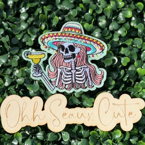 May include: Embroidered patch featuring a skeleton wearing a sombrero and holding a cocktail. The skeleton has pink hair and is on a light green background. Below is a wooden cutout with the words "Ohh Seaux Cute".