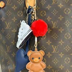 May include: A brown leather-like teddy bear charm, a white miniature sneaker charm, a dark blue heart-shaped charm, and a bright red fluffy pom-pom attached to a gold-toned metal clasp hanging from a brown monogrammed tote bag.