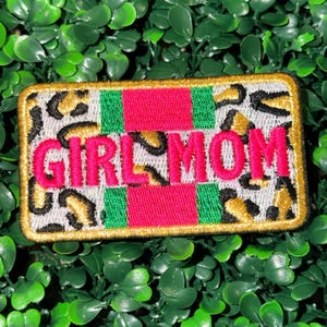 May include: Embroidered patch with the words "GIRL MOM" in bright pink. The patch has a leopard print background with red and green stripes. The border is gold.