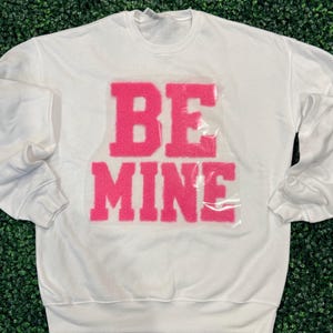 May include: A white sweatshirt with the words "BE MINE" in bright pink, textured letters across the front. The sweatshirt has long sleeves and a crew neck. The background is a green, textured surface.