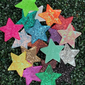 May include: A collection of colorful star-shaped sequin patches in various colors, including pink, green, orange, and gold. The patches are arranged on a green, textured background. These decorative patches are ideal for embellishing clothing or accessories.