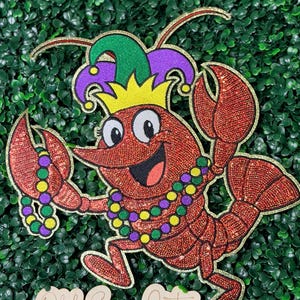 May include: A glittery red crawfish cartoon character wearing a jester hat and a beaded necklace. The crawfish has a smiling face and is holding its claws up. The text "Ohh Seaux Cute" is at the bottom.