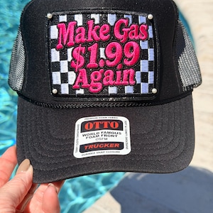 May include: A black trucker hat with a mesh back and a rectangular patch featuring a black and white checkered pattern. The patch displays the pink embroidered text "Make Gas $1.99 Again" and has small white beads at each corner. An Otto brand sticker is on the brim.