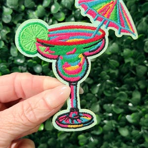 May include: Embroidered patch featuring a colorful margarita cocktail with a lime wedge and a miniature umbrella. The patch has a vibrant design with shades of pink, green, yellow, and blue, outlined in black. The patch is held against a green background.
