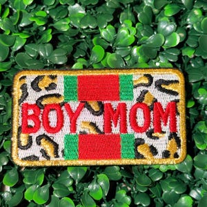 May include: Rectangular patch with the text "BOY MOM" in red, set against a leopard print background. The patch has red and green stripes and a gold border. The patch is on a green leafy background.
