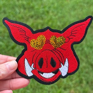 May include: Embroidered patch featuring a red warthog face with black outlines. The warthog has gold sequin heart-shaped eyes, white tusks, and a smiling mouth. The patch is held against a green background.