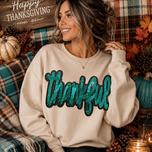 May include: A beige crewneck sweatshirt featuring the word Thankful in large, cursive, emerald green sequined lettering with a black outline. The long-sleeved pullover has a ribbed neckline, cuffs, and hem, providing a relaxed fit for the wearer.
