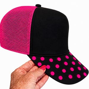 May include: A baseball cap featuring a solid black front panel and a vibrant hot pink mesh back. The curved brim is black and decorated with a repeating pattern of bright hot pink polka dots. The cap includes a small black button on the top center.
