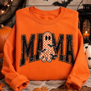 May include: An orange long-sleeve sweatshirt featuring the word MAMA in large black block letters with a glittery texture. The letter A is replaced by a ghost with an orange and white checkered pattern, accented by small black bat silhouettes.