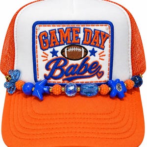May include: An orange and white trucker hat featuring a rectangular patch with the text "GAME DAY Babe" in blue and orange embroidery, centered above a football graphic. A strand of blue, orange, and clear beads decorates the front brim base.