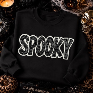 May include: A black long-sleeve sweatshirt featuring the word SPOOKY in large, black sequined letters with a white border. The text is centered on the chest. The garment has a crew neckline and ribbed cuffs, providing a simple, clean design.