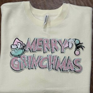 May include: Cream-colored sweatshirt with the text "MERRY GRINCHMAS" in pink and silver sequins. The design includes Grinch-themed illustrations in shades of pink, green, and white. A festive holiday-themed apparel item.