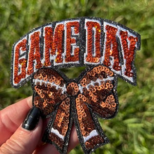 May include: A sequined patch with the text "GAME DAY" in orange letters on a black background. Below the text is a bow with orange sequins and white accents. The patch is held against a blurred green background.