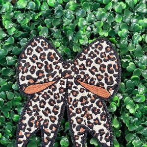 May include: Embroidered bow with a leopard print pattern in shades of brown, black, and cream. The bow has a brown center and black trim, set against a backdrop of green foliage.