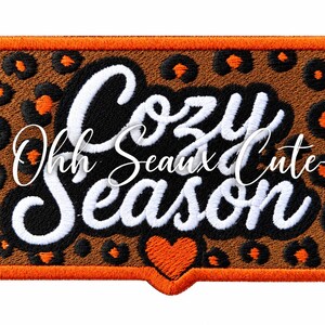 May include: A rectangular embroidered patch featuring a brown and black leopard print background with an orange border. The text "Cozy Season" is stitched in white, and a small orange heart is positioned at the bottom center of the patch.