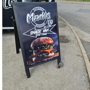 May include: Black A-frame sign advertising "Gourmet Munchies Burgers" and "Snack Van Food & Drinks." The sign features a photo of a large burger with sesame seed bun, cheese, lettuce, and tomato. The sign is on a sidewalk.