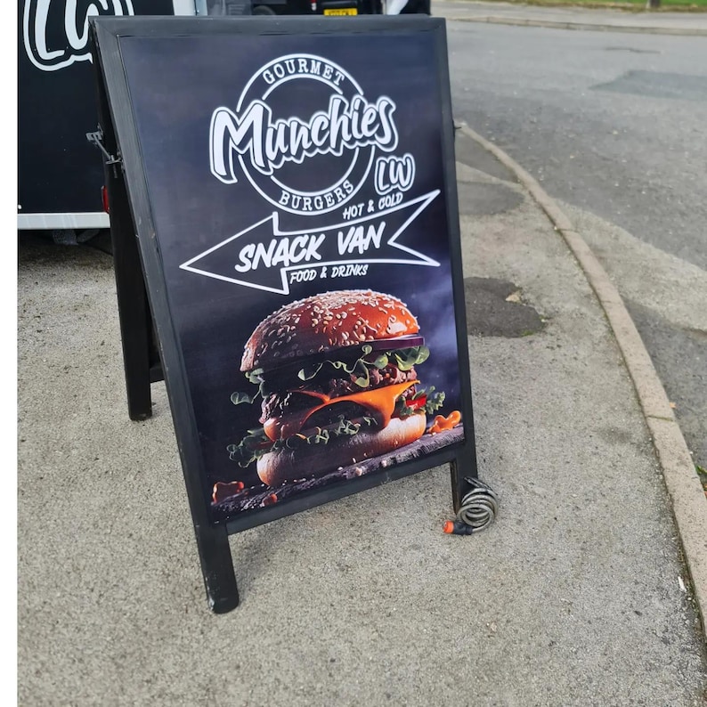 May include: Black A-frame sign advertising "Gourmet Munchies Burgers" and "Snack Van Food & Drinks." The sign features a photo of a large burger with sesame seed bun, cheese, lettuce, and tomato. The sign is on a sidewalk.
