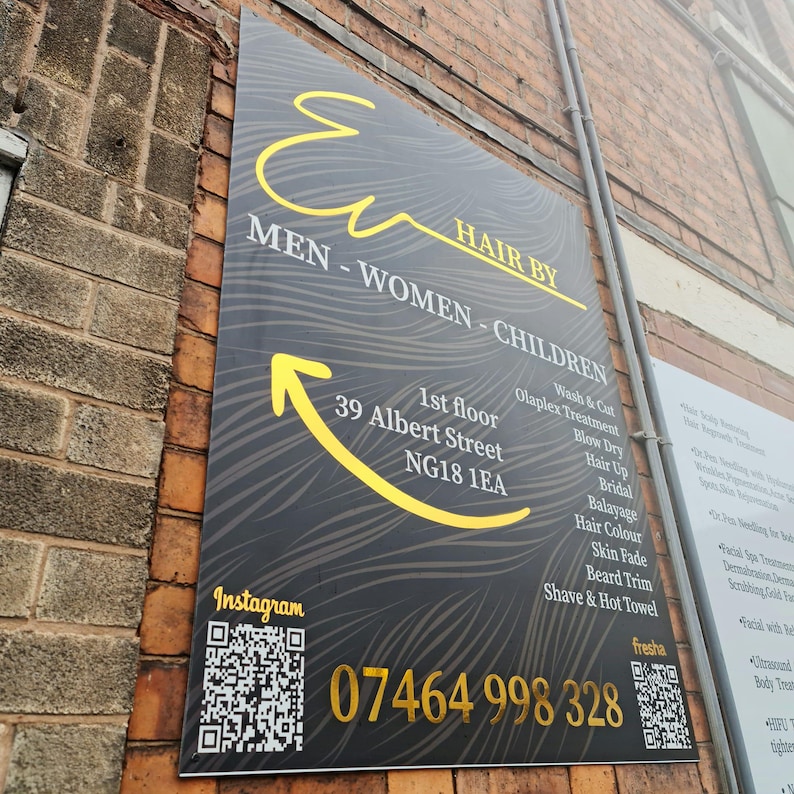 May include: A rectangular sign for a hair salon, mounted on a brick wall. The sign is black with yellow text and a yellow arrow. It reads "HAIR BY" and lists services like "Wash & Cut" and "Skin Fade". The phone number 07464 998 328 is displayed.