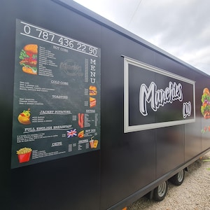 May include: A black food truck with the name "Munchies" in white script. The truck has a menu board with food illustrations and a phone number. The menu includes hot dogs, cold subs, toasties, jacket potatoes, and a full English breakfast.