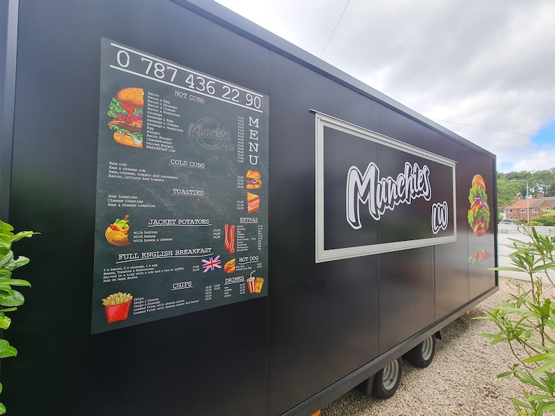 May include: A black food truck with the name "Munchies" in white script. The truck has a menu board with food illustrations and a phone number. The menu includes hot dogs, cold subs, toasties, jacket potatoes, and a full English breakfast.