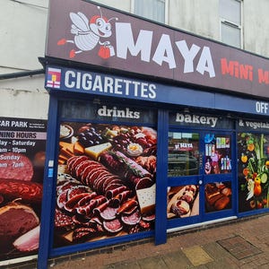 May include: Exterior shot of a small convenience store called Maya Mini Market. The store has a blue awning with the words "Cigarettes", "Drinks", "Bakery", "& Vegetables", and "Off Licence". A sign advertises a free car park and opening hours. The store's windows display images of food.