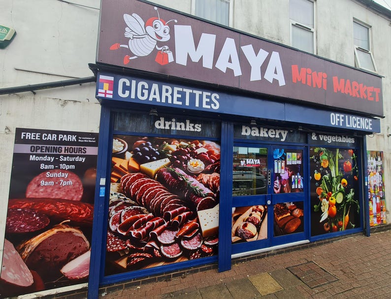 May include: Exterior shot of a small convenience store called Maya Mini Market. The store has a blue awning with the words "Cigarettes", "Drinks", "Bakery", "& Vegetables", and "Off Licence". A sign advertises a free car park and opening hours. The store's windows display images of food.