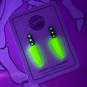 May include: Two glow-in-the-dark knife earrings on a white card. The earrings feature black handles with green dots and glowing green blades. The card displays the text "Creepy Clayworks" and a website address.